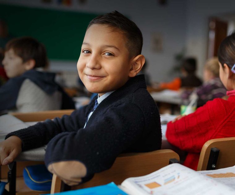 Menino sorridente sentado em sala de aula, olhando para a câmera, com livros abertos à sua frente. A imagem ilustra os resultados positivos da experiência de Sobral na educação.