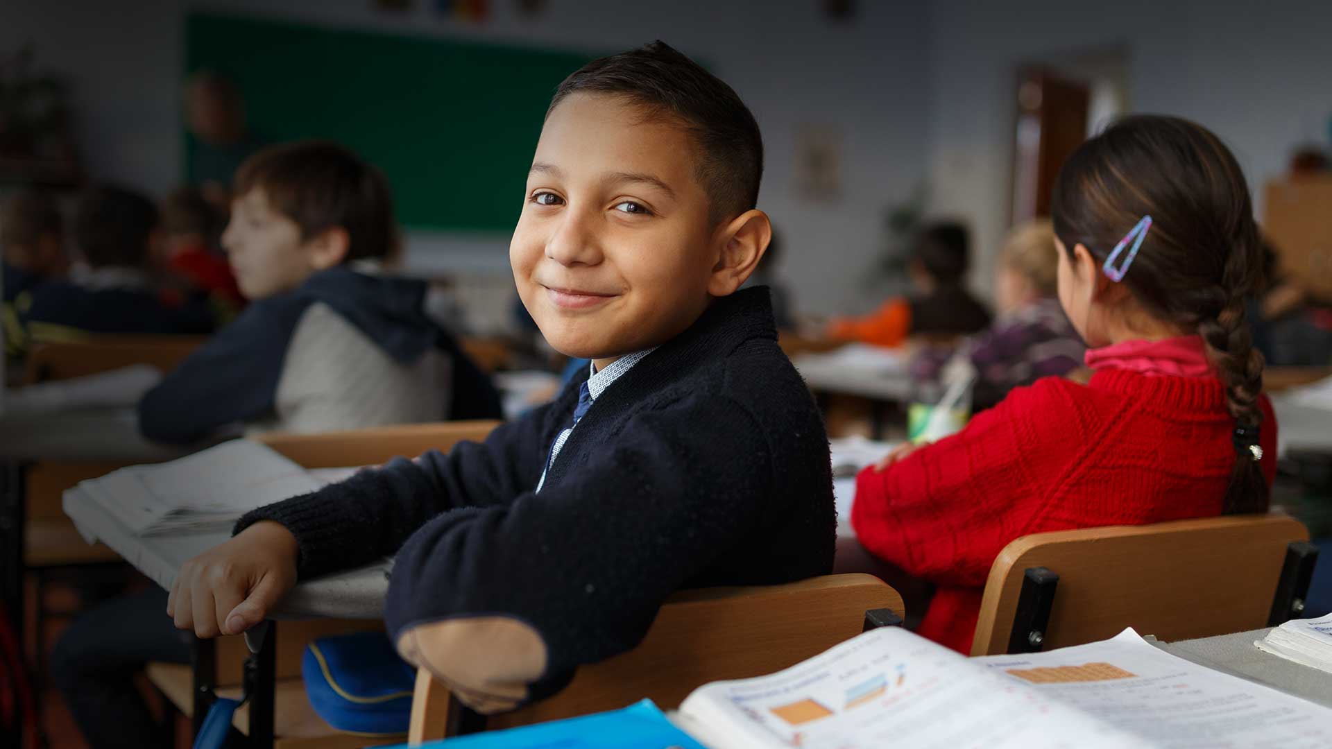 Menino sorridente sentado em sala de aula, olhando para a câmera, com livros abertos à sua frente. A imagem ilustra os resultados positivos da experiência de Sobral na educação.