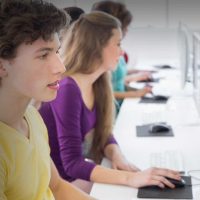 A imagem sugere a aplicação da prova do PISA 2025 em uma sala de aula com alguns estudantes com seus notebooks e uma professora.