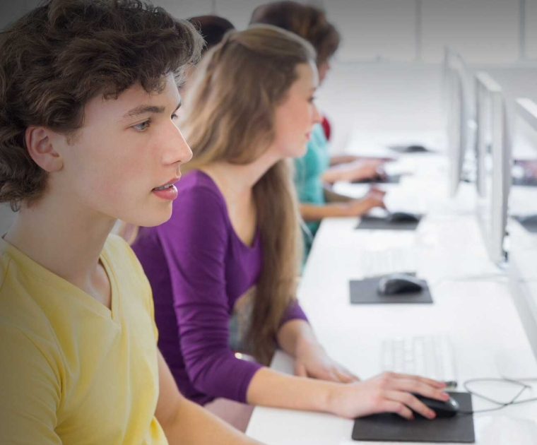 A imagem sugere a aplicação da prova do PISA 2025 em uma sala de aula com alguns estudantes com seus notebooks e uma professora.